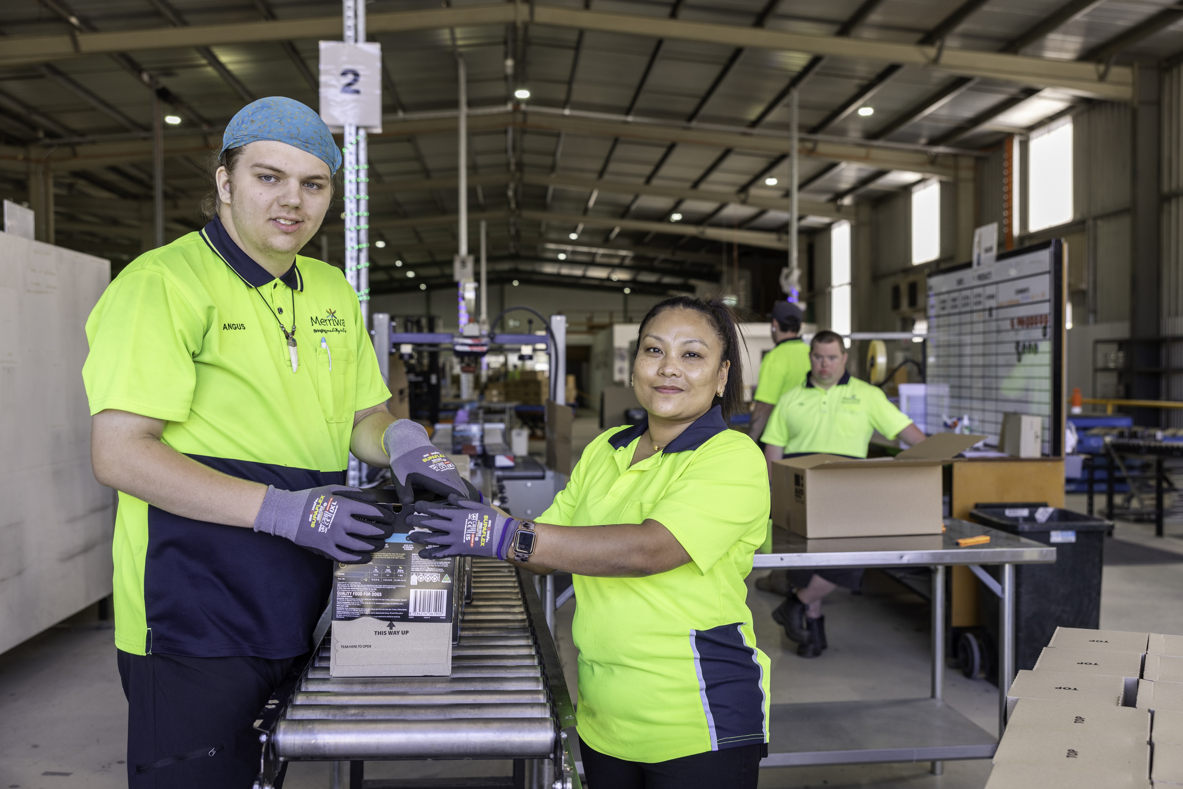 Two workers in hi-vis shirts pack products on a conveyor line inside a warehouse, smiling toward the camera.
